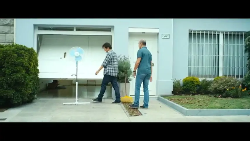 Two men stand in front of a house, one holding a broom and the other walking towards him, then they both stand talking next to a fan in the garage.