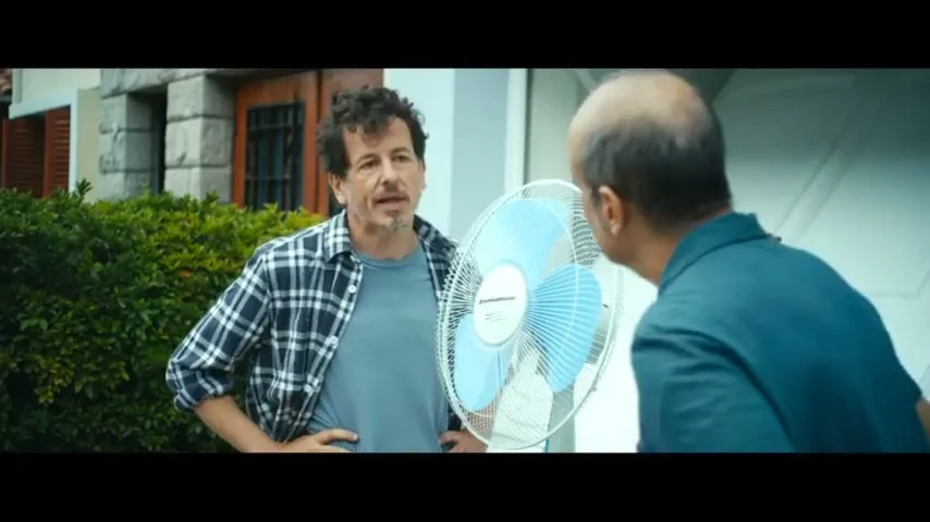 Two men stand outside a house, one holding a blue fan, talking to each other in a casual conversation.