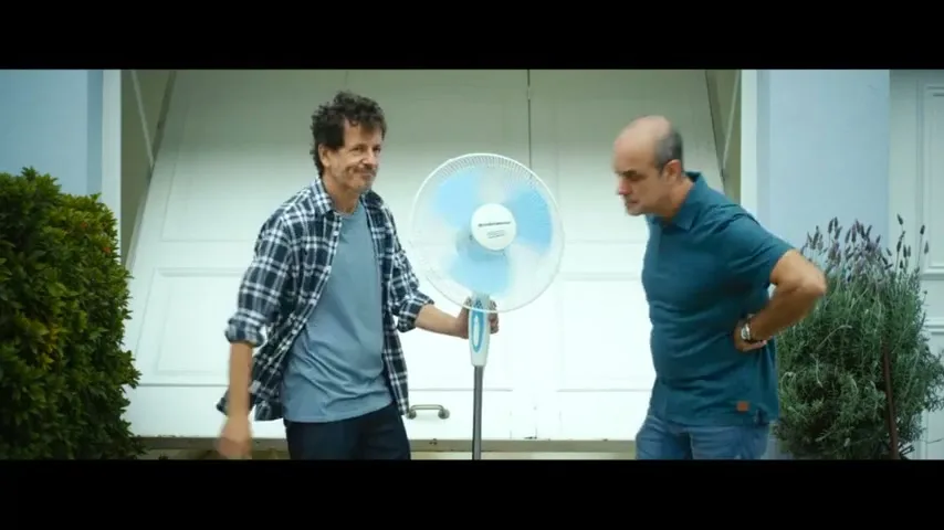 Two men stand outside a garage, one holding a fan while the other leans in to feel the air, both smiling.