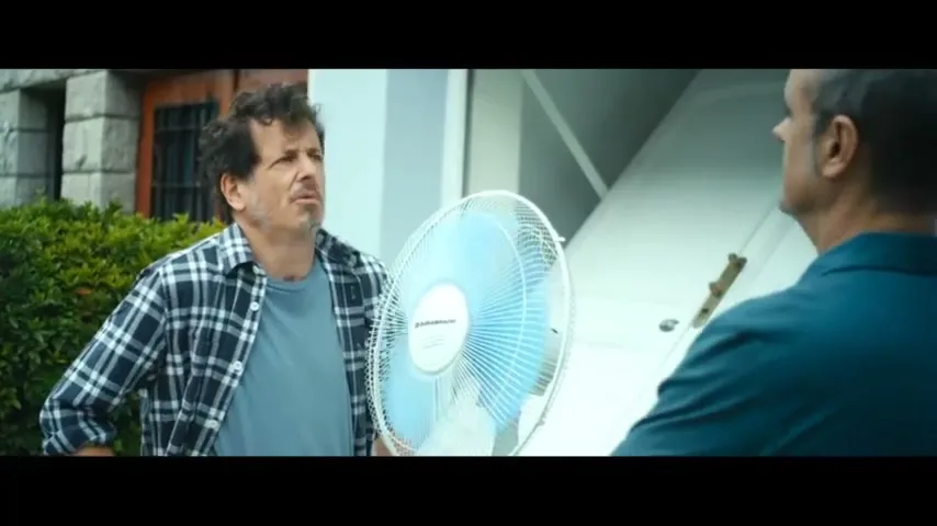 A man in a plaid shirt holds a fan and talks to another man, whose back is to the camera, in an outdoor setting.