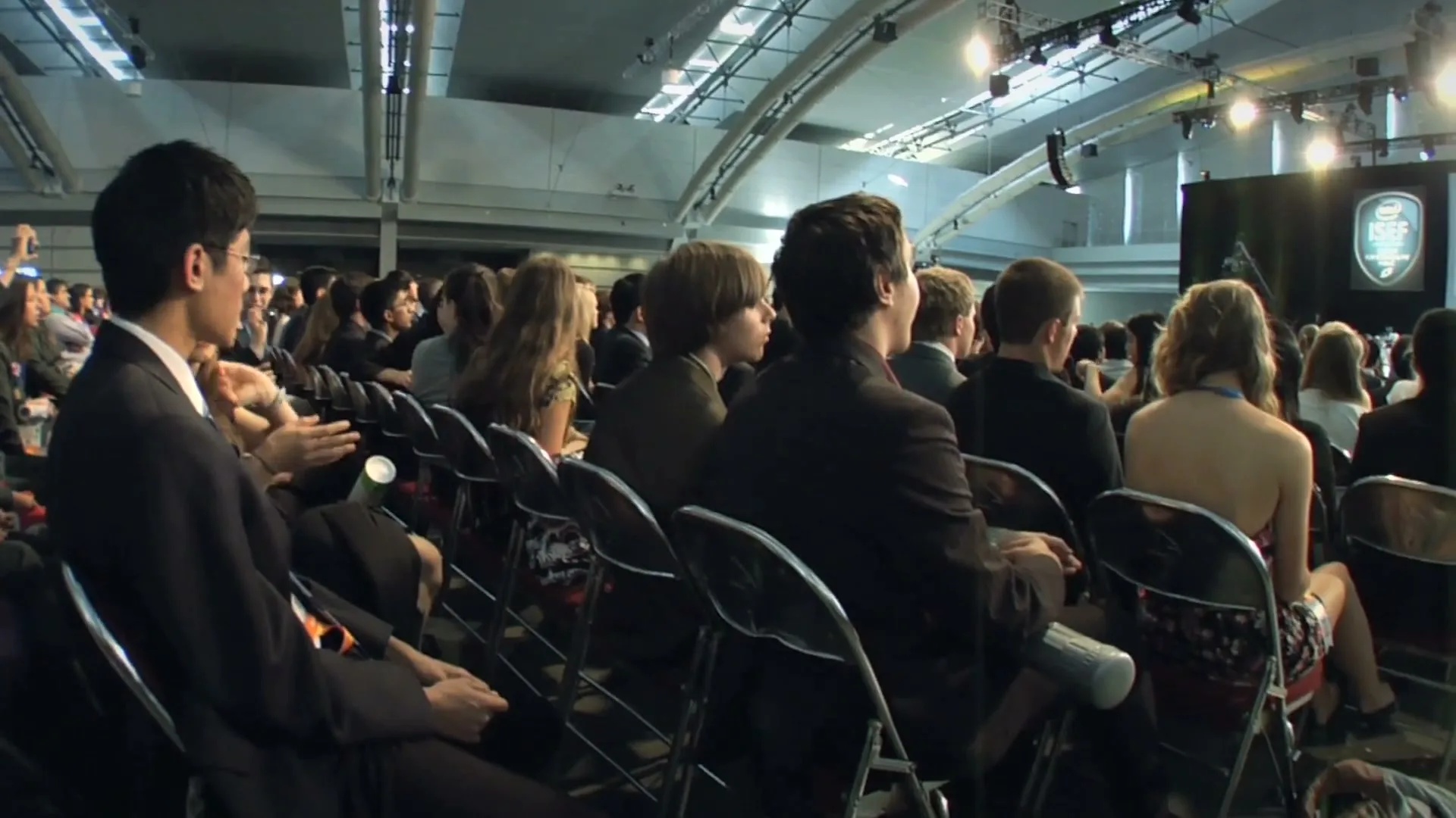 A diverse audience of young people in formal attire sits in rows of chairs, looking towards a brightly lit stage at a large indoor event.