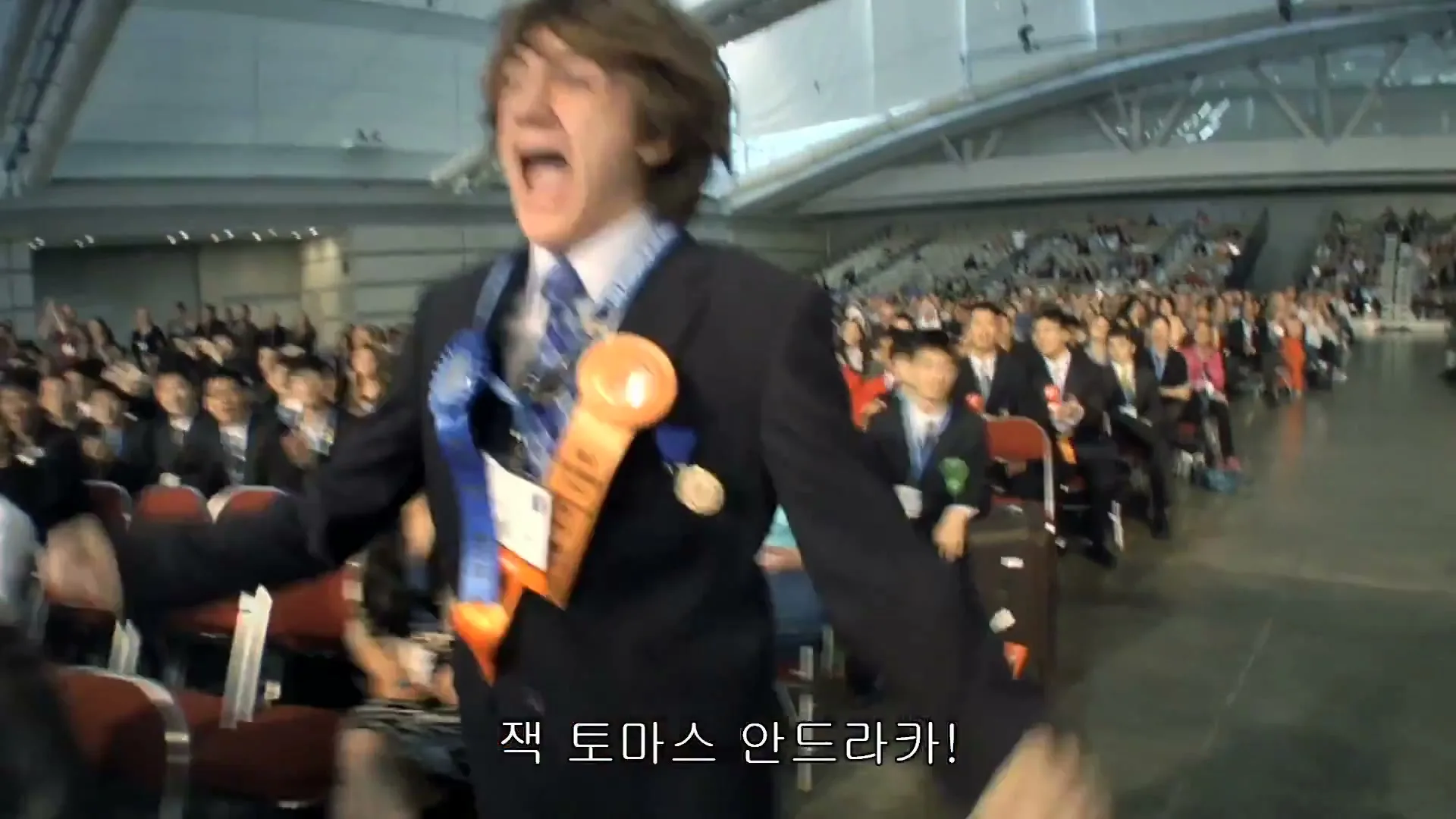 A young male student, wearing a suit and ribbons, stands up and cheers excitedly in a large auditorium filled with clapping students.
