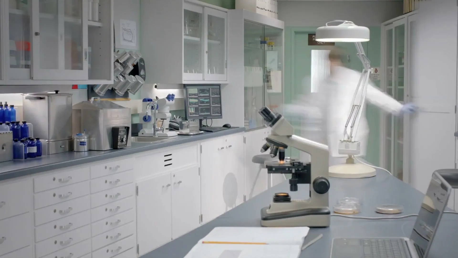 A young male scientist in a lab coat works at a computer and microscope in a brightly lit laboratory, then quickly exits the frame.