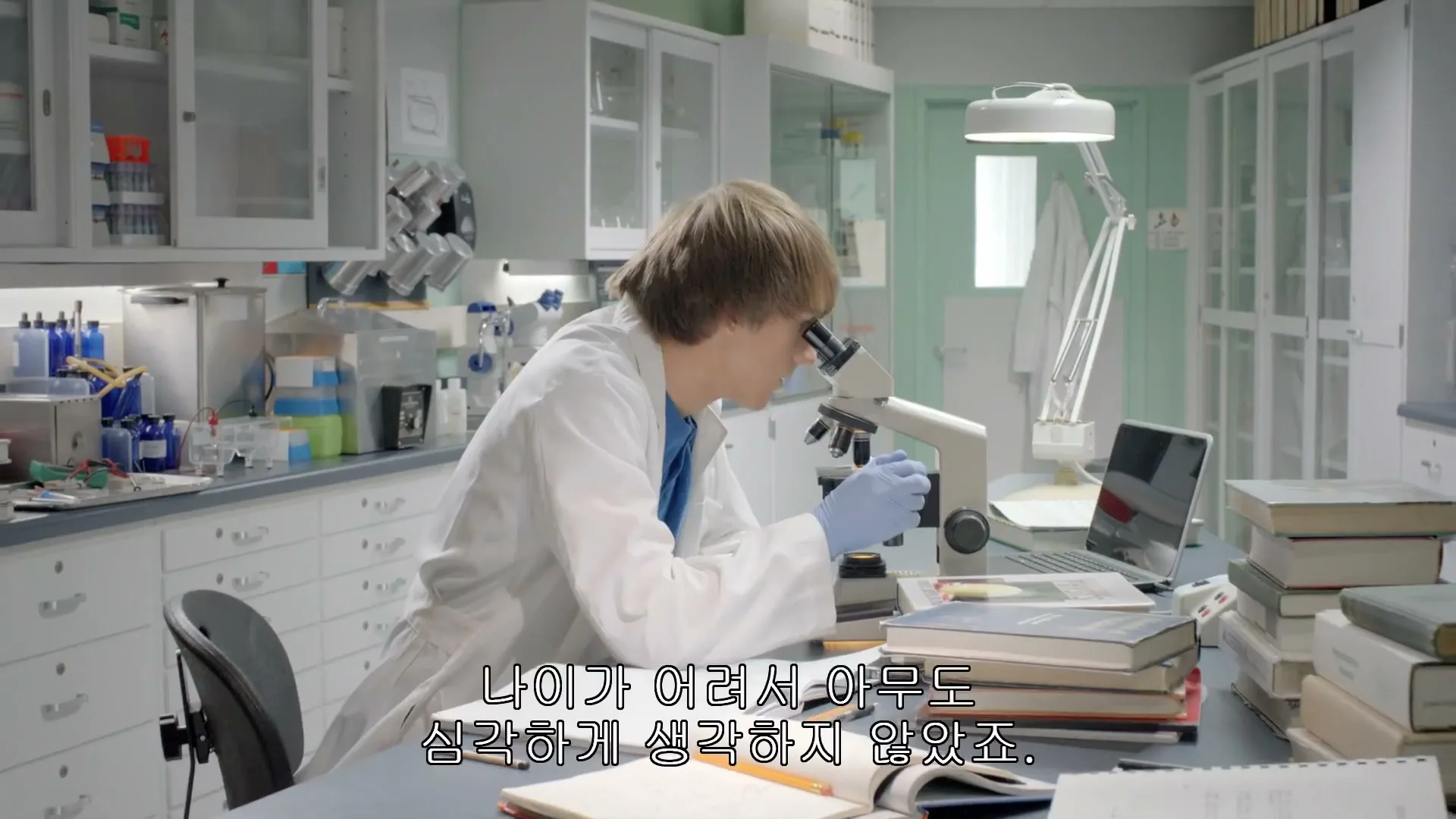 A young male scientist in a lab coat and blue gloves looks into a microscope, then looks up in frustration in a brightly lit laboratory.