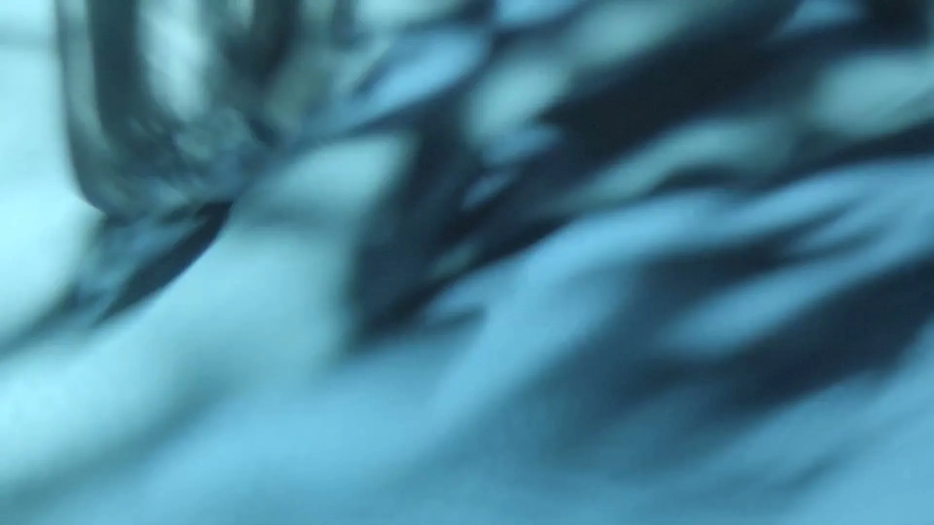 An extreme close-up shot of a blurry, abstract blue surface with dark shadows, possibly water or a reflective material, creating a mysterious and fluid visual.