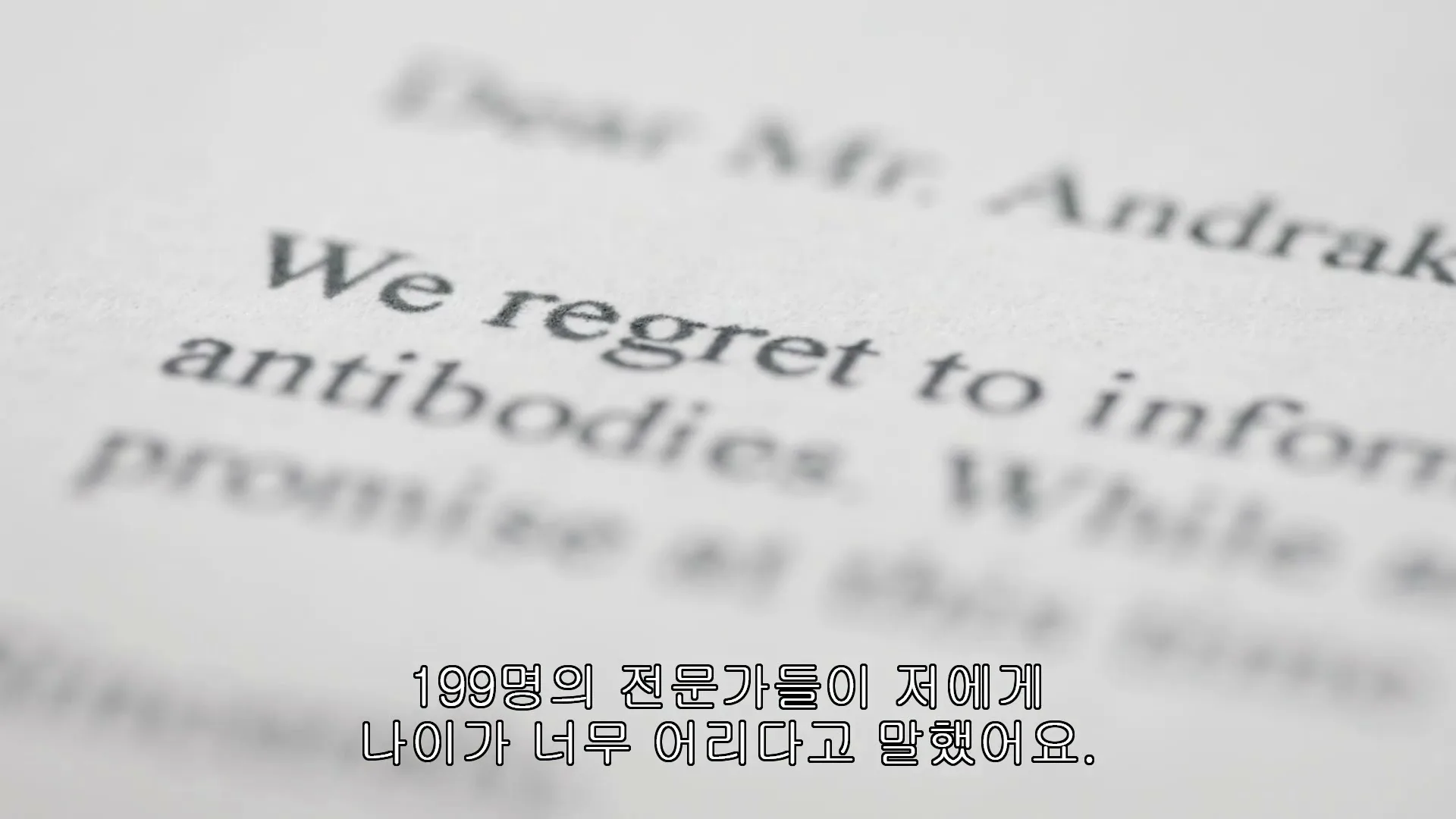 A close-up shot of a rejection letter with the words "Dear Mr. Andraka" and "We regret to inform" visible, with Korean subtitles at the bottom.