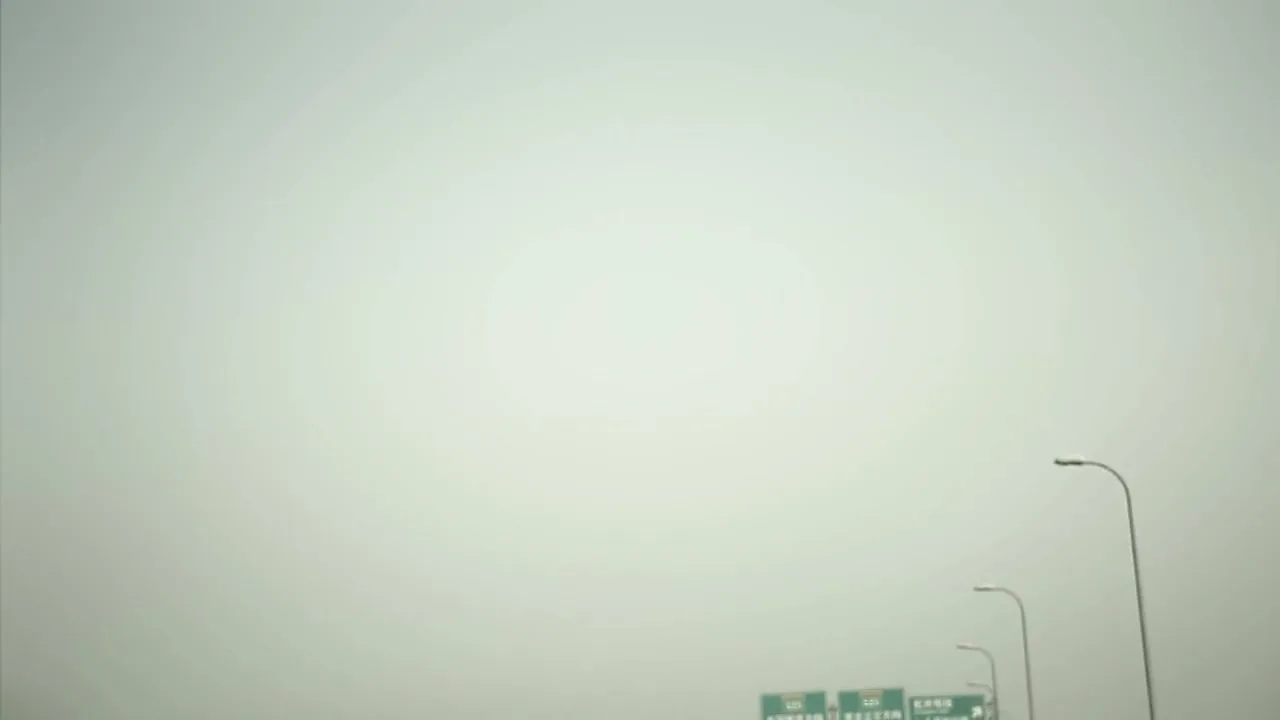 A wide shot of a highway with streetlights and a hazy, overcast sky, as a car drives forward.