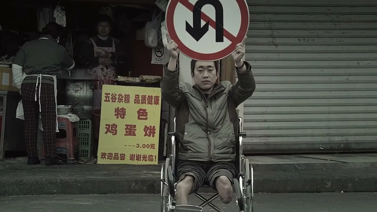 A man in a wheelchair, missing both legs, holds up a "no U-turn" sign on a street in front of a small shop.