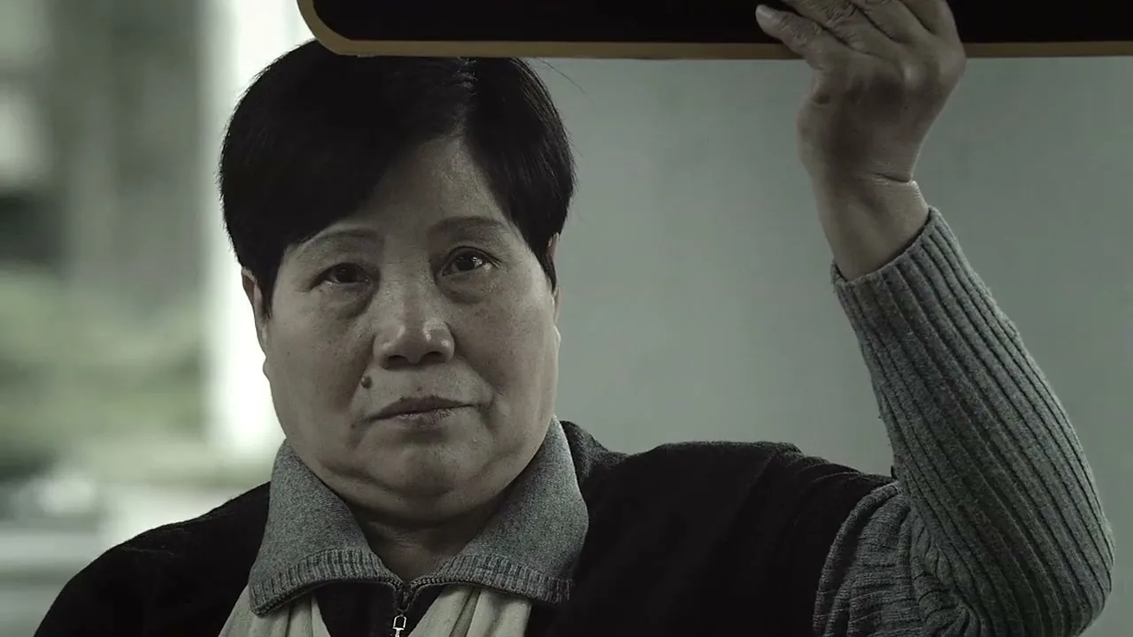A middle-aged Asian woman with short dark hair, wearing a dark jacket and light-colored shirt, holds a dark sign above her head, looking directly at the camera in a medium shot.
