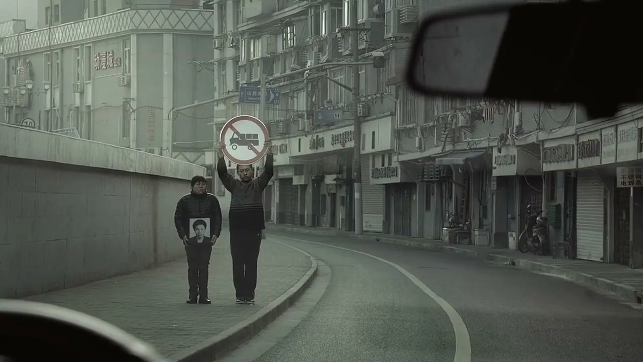 Two men stand on a sidewalk, one holding a "no trucks" sign above his head, the other holding a framed photo, on a city street.