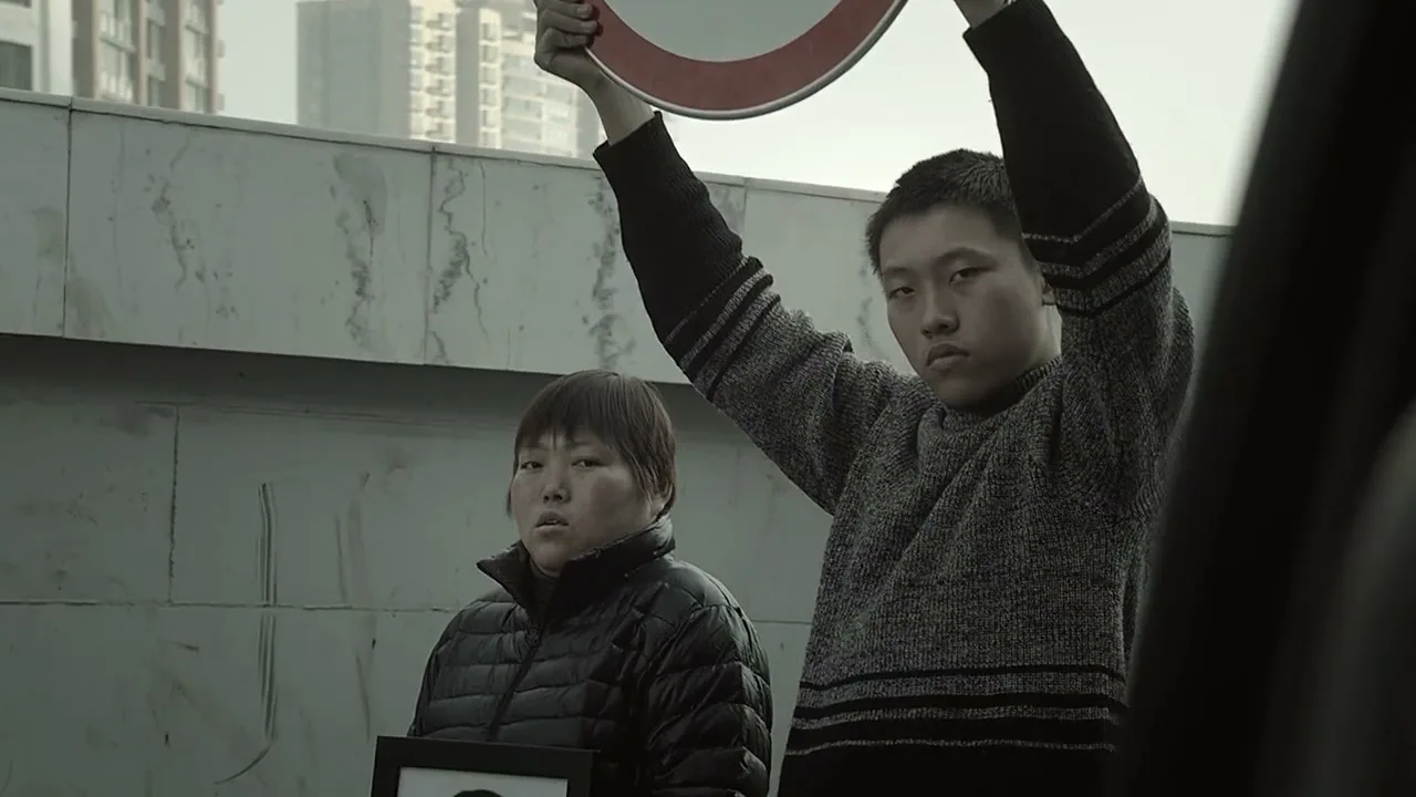 Two young people, a male and a female, stand on a concrete overpass, holding up a traffic sign and a framed photo, looking serious.