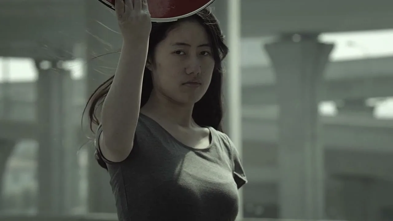 A young Asian woman in a grey t-shirt holds a red traffic sign above her head, looking seriously at the camera on a highway overpass.