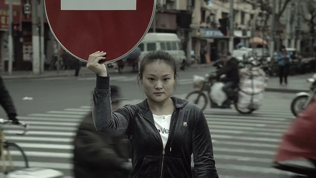 A young woman with a serious expression holds a red and white stop sign above her head, standing in the middle of a busy street with traffic and buildings in the background.