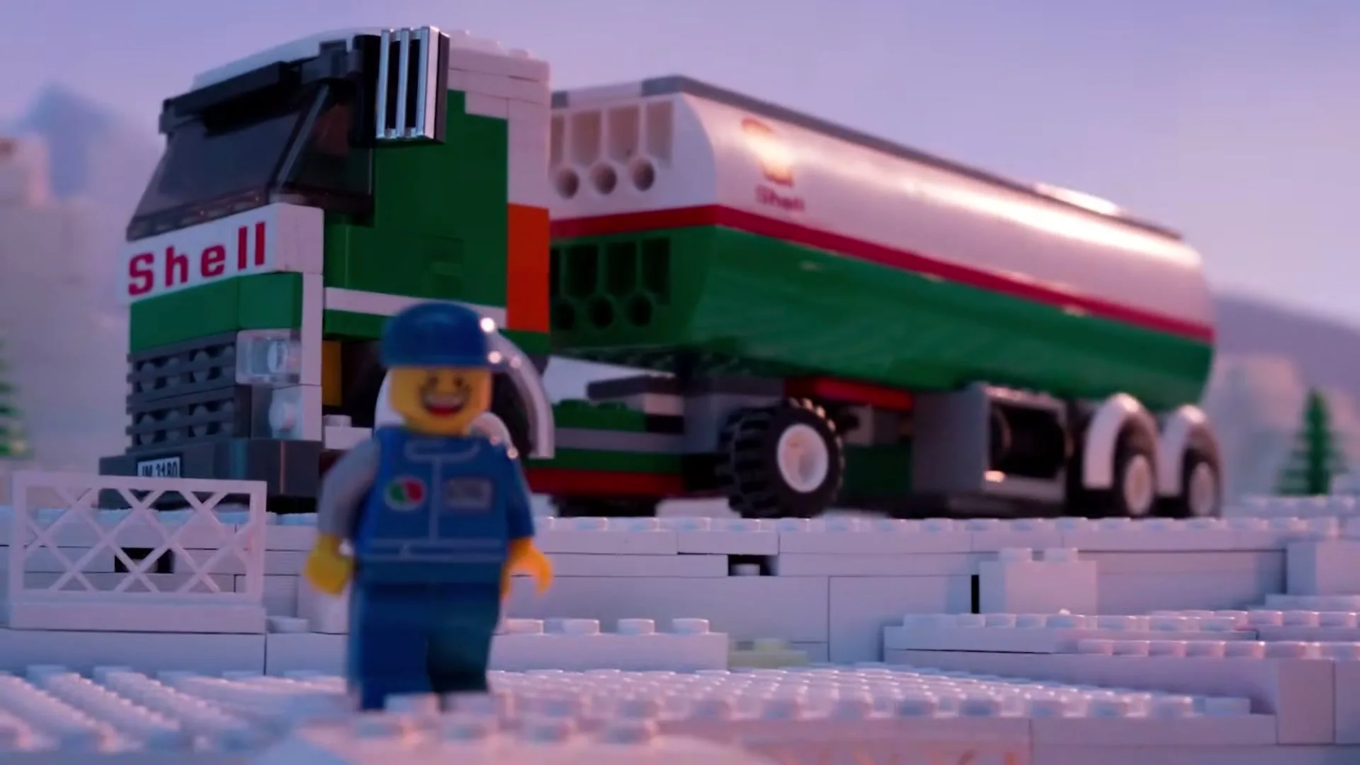 A Lego oil worker minifigure stands in a snowy landscape as a Shell oil tanker truck drives past in the background.