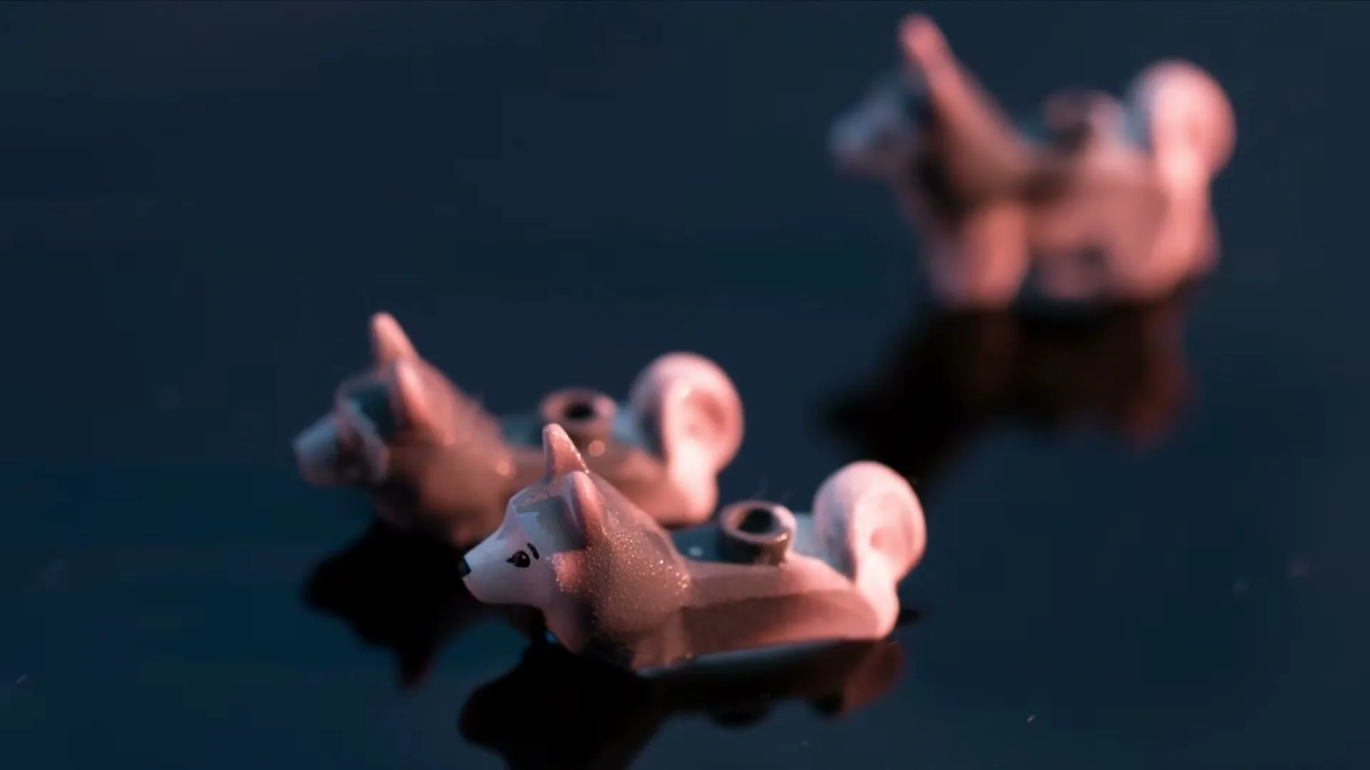 Three small, grey and white Lego husky dog figures are partially submerged in dark water, with a pinkish light reflecting on them.