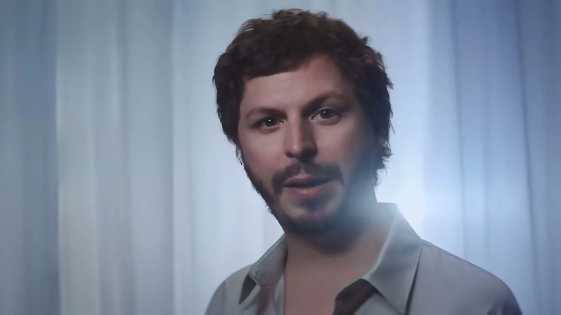 Michael Cera, a man with dark curly hair and a beard, holds up a jar of CeraVe Moisturizing Cream, looking at it with a slight smile in a studio.