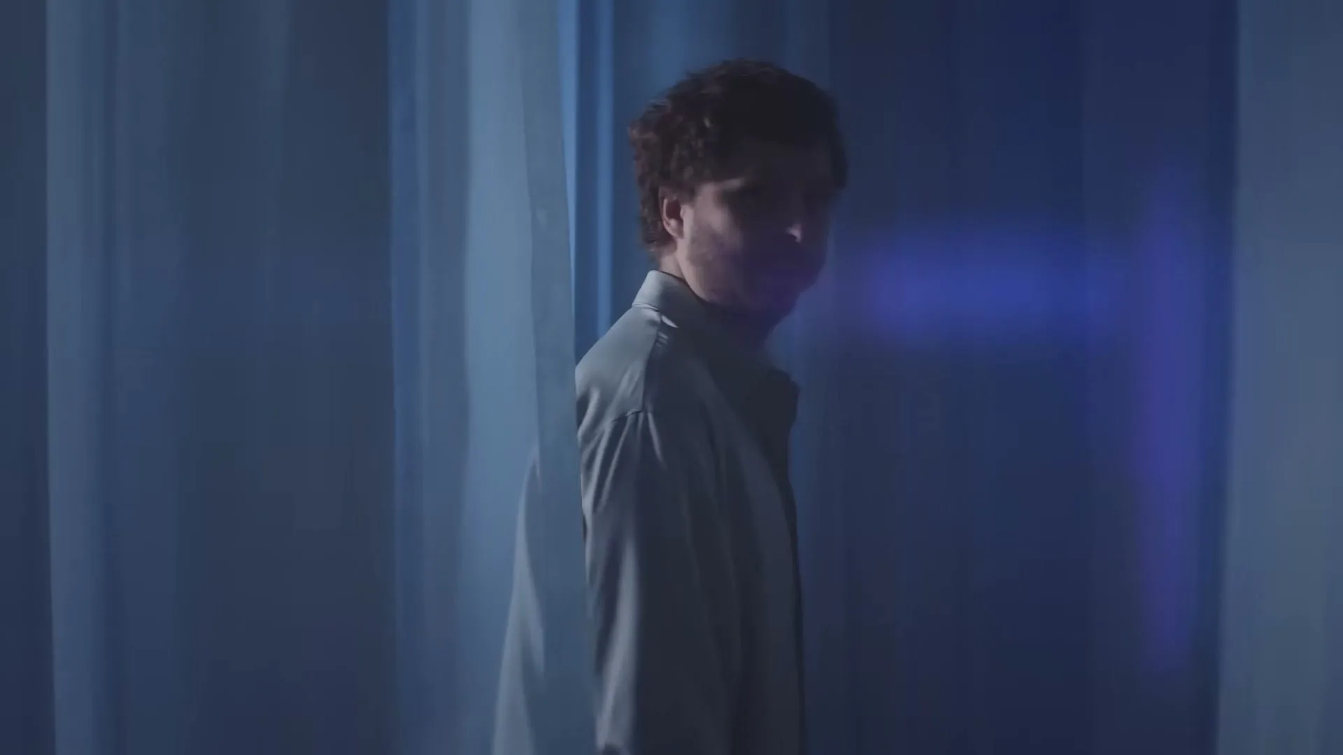 Michael Cera, a man with dark curly hair and a beard, holds a CeraVe Moisturizing Cream jar and looks at it, then turns to look over his shoulder in a blue-lit room.