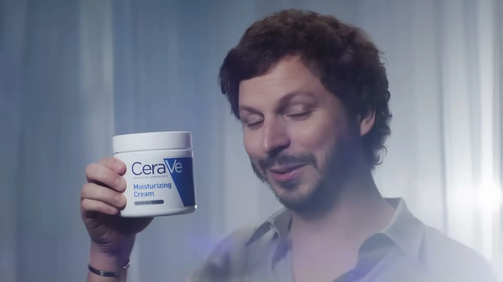 Michael Cera, a man with curly brown hair and a mustache, holds up a white jar of CeraVe Moisturizing Cream, looking at it with a slight smile.