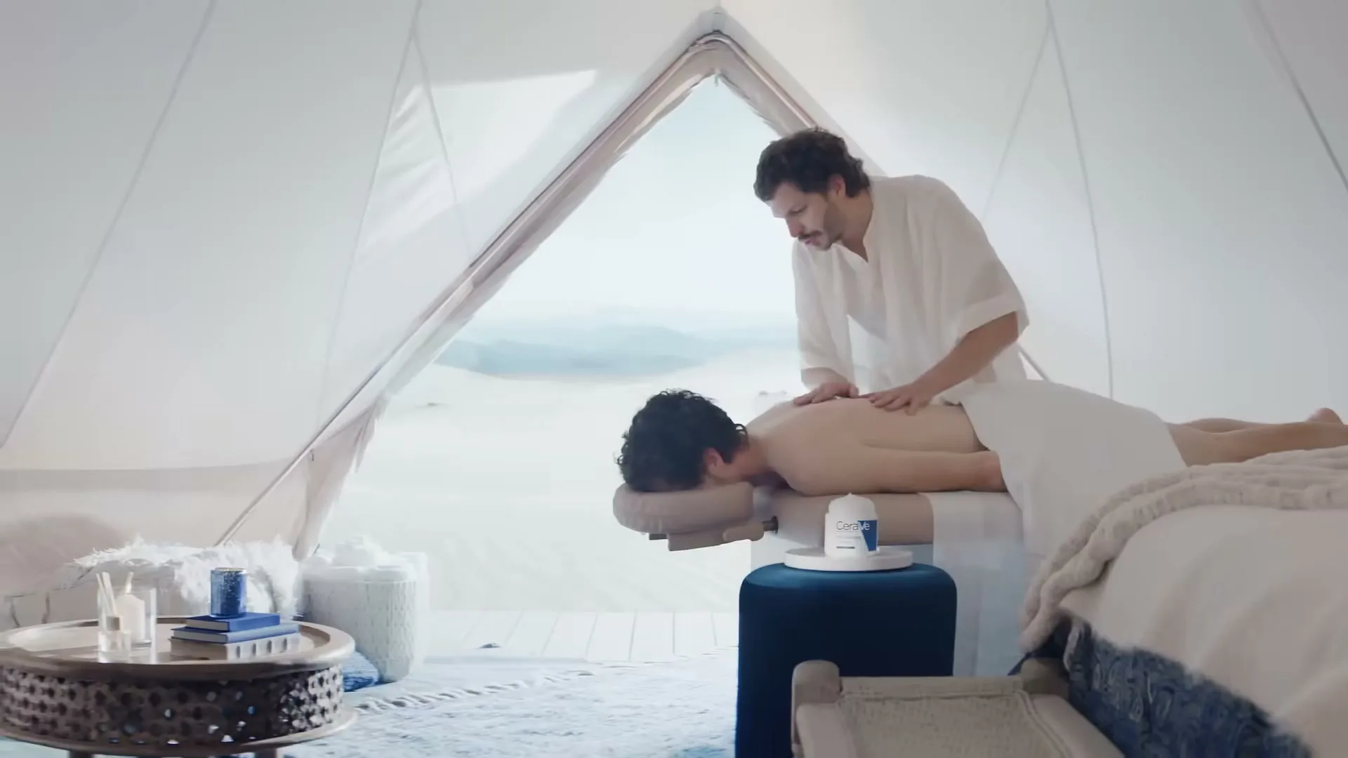 A man with curly hair receives a back massage from another man in a white tent, with a desert landscape visible outside.