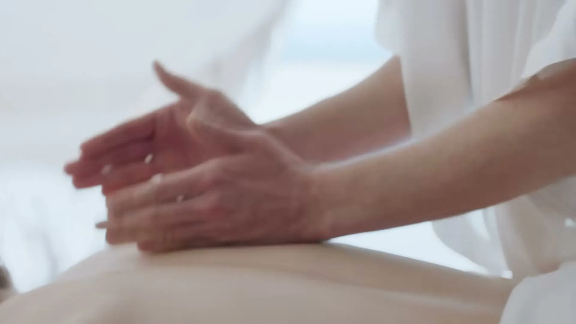 A person with pale skin lies face down as another person's hands massage their back in a brightly lit, serene spa setting.