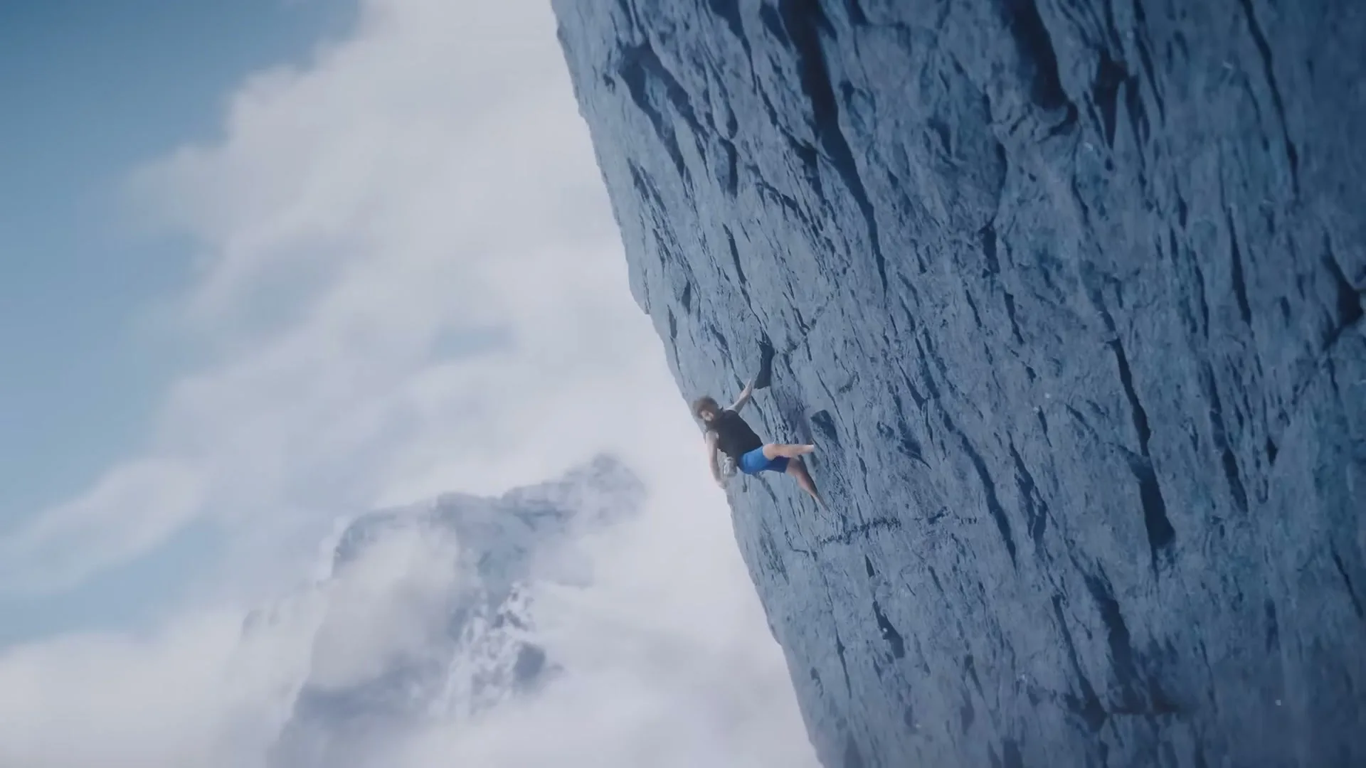 A man in a black shirt and blue shorts is rock climbing up a sheer cliff face, with clouds and mountains in the background.