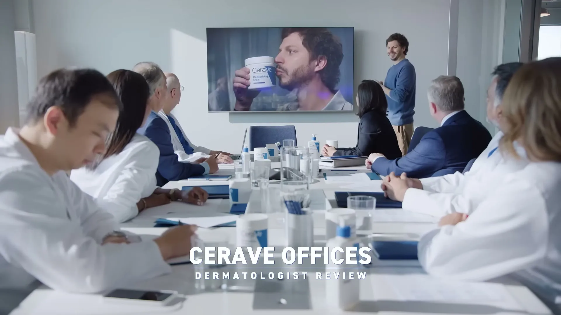 A group of doctors and business people sit around a conference table, watching a man on a large screen holding a CeraVe product, while another man stands by the screen.