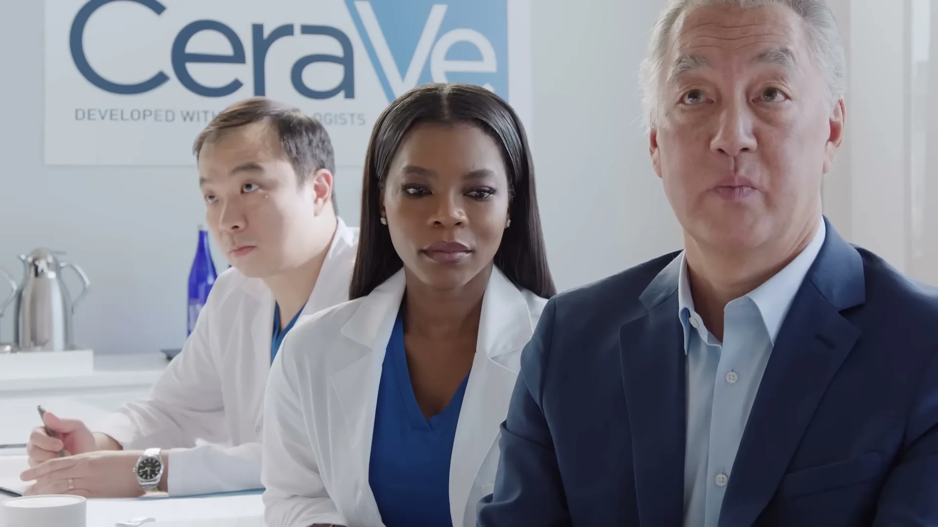 Three people, two doctors and one man in a suit, sit in a meeting room with the CeraVe logo on the wall, looking directly at the camera.