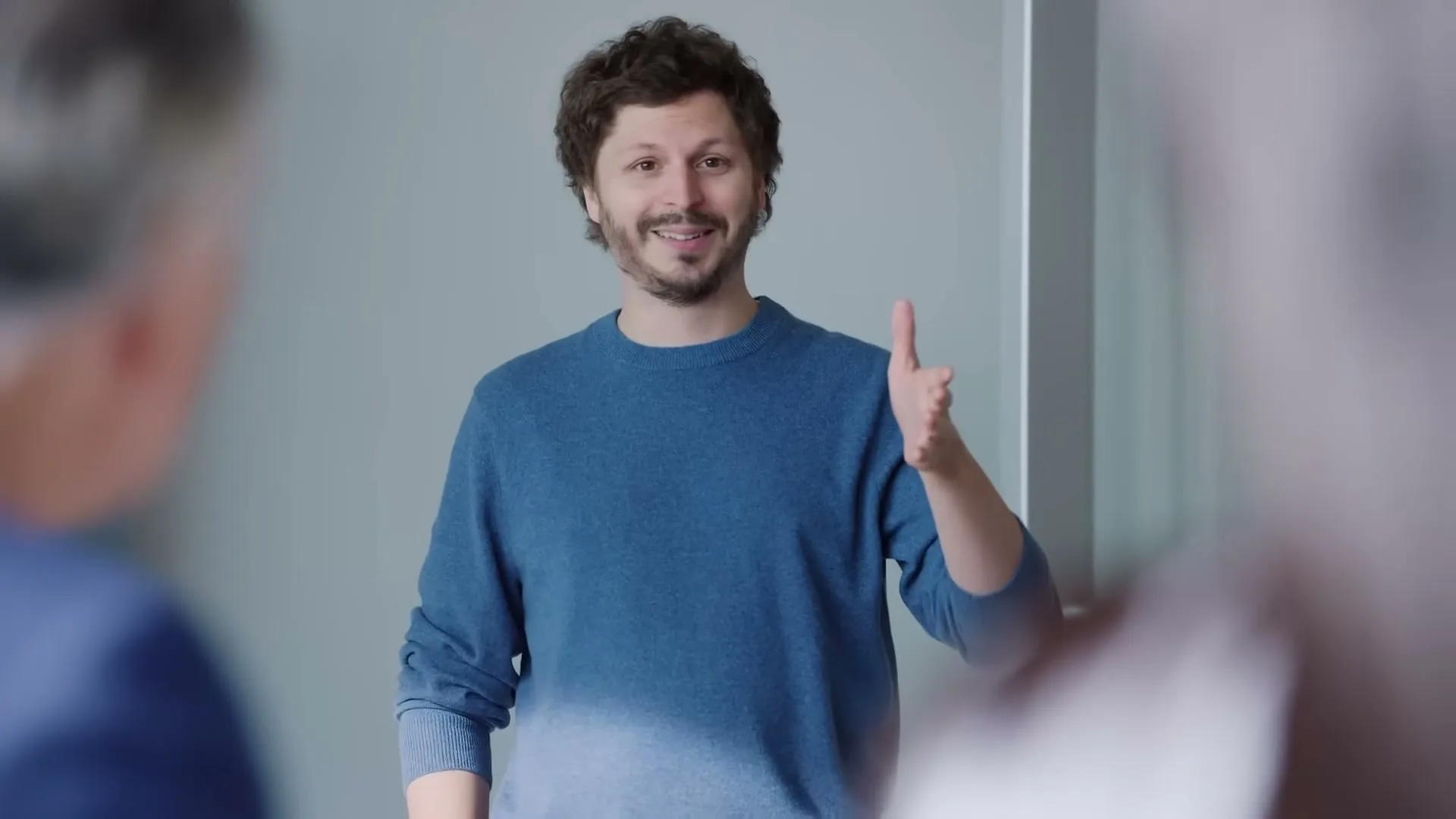Michael Cera, a male in his 30s with a beard, wearing a blue sweater, stands in a bright room, gesturing with his hands while talking to two people whose backs are to the camera.