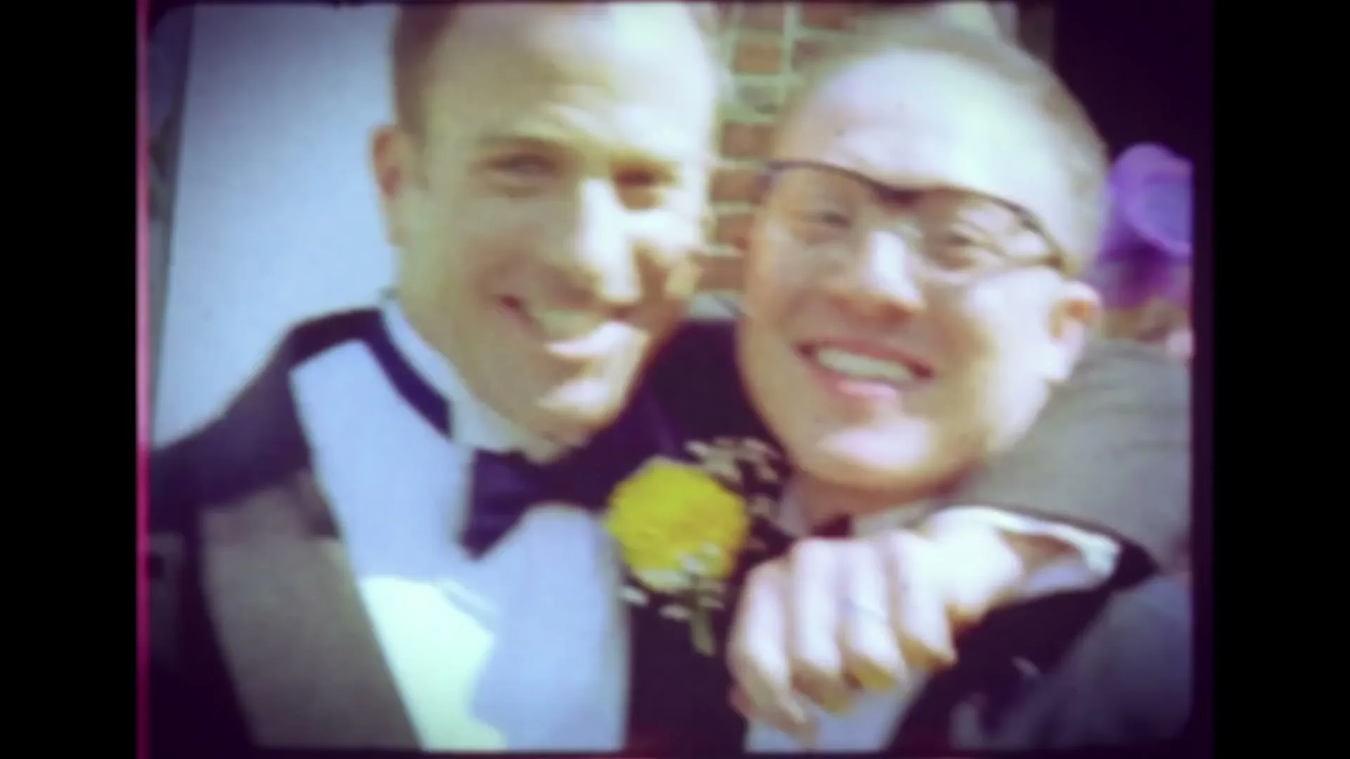 A vintage home video shows two men in tuxedos smiling and embracing, followed by two women in wedding dresses laughing and posing for the camera.