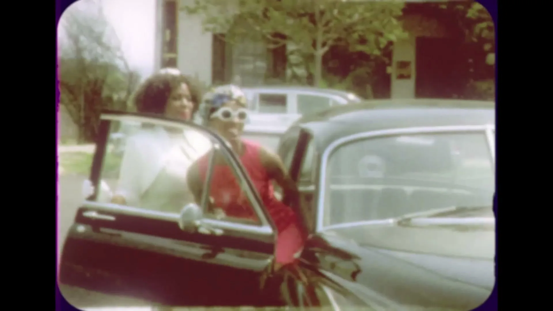 Two women, one in a white dress and the other in a pink dress and headscarf, are getting into a black car on a sunny day in a suburban setting.