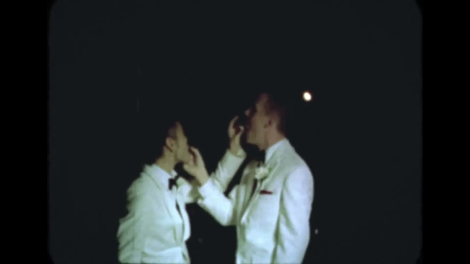 Two men in white tuxedos feed each other cake at a wedding reception at night.