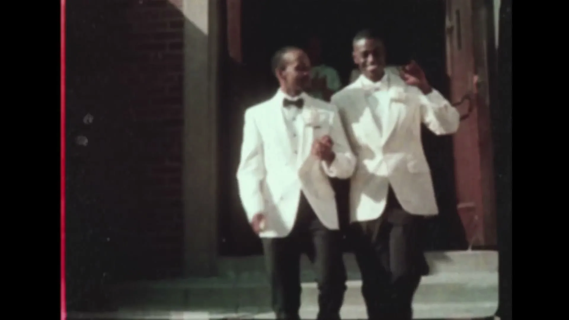 Two young Black men in white tuxedos and black pants walk down steps from a building entrance, smiling and waving.