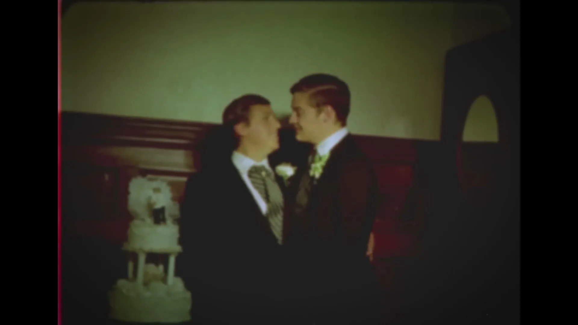 Two men in suits and ties, one with a boutonniere, stand next to a wedding cake and kiss at their wedding reception.