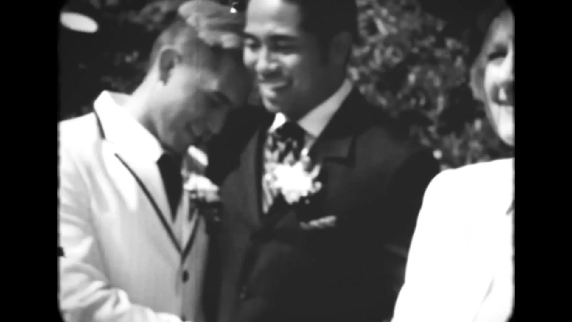 Two men in suits, one in white and one in black, smile and laugh together at an outdoor event, with a woman partially visible on the right.