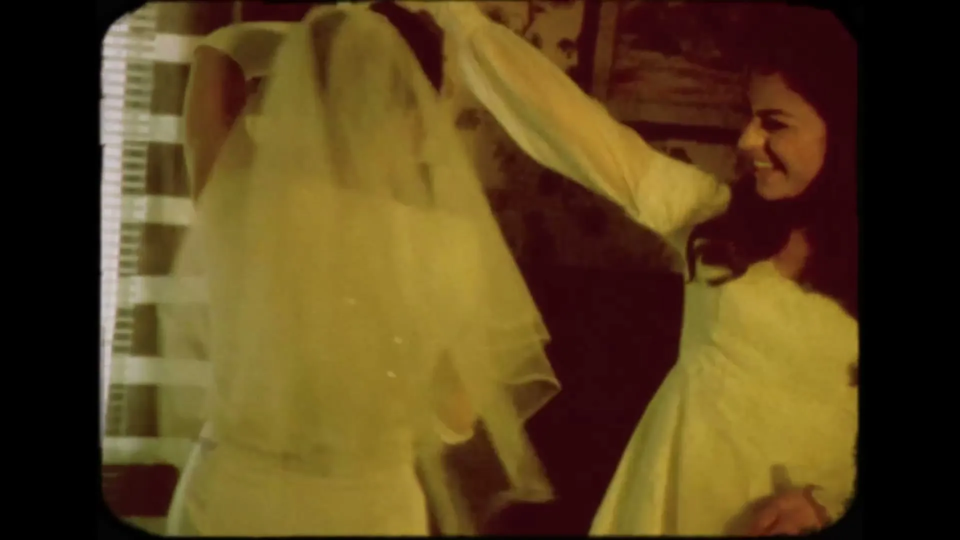 Two women, one in a wedding dress and veil, the other in a white dress, are laughing and dancing together in a room with striped curtains.