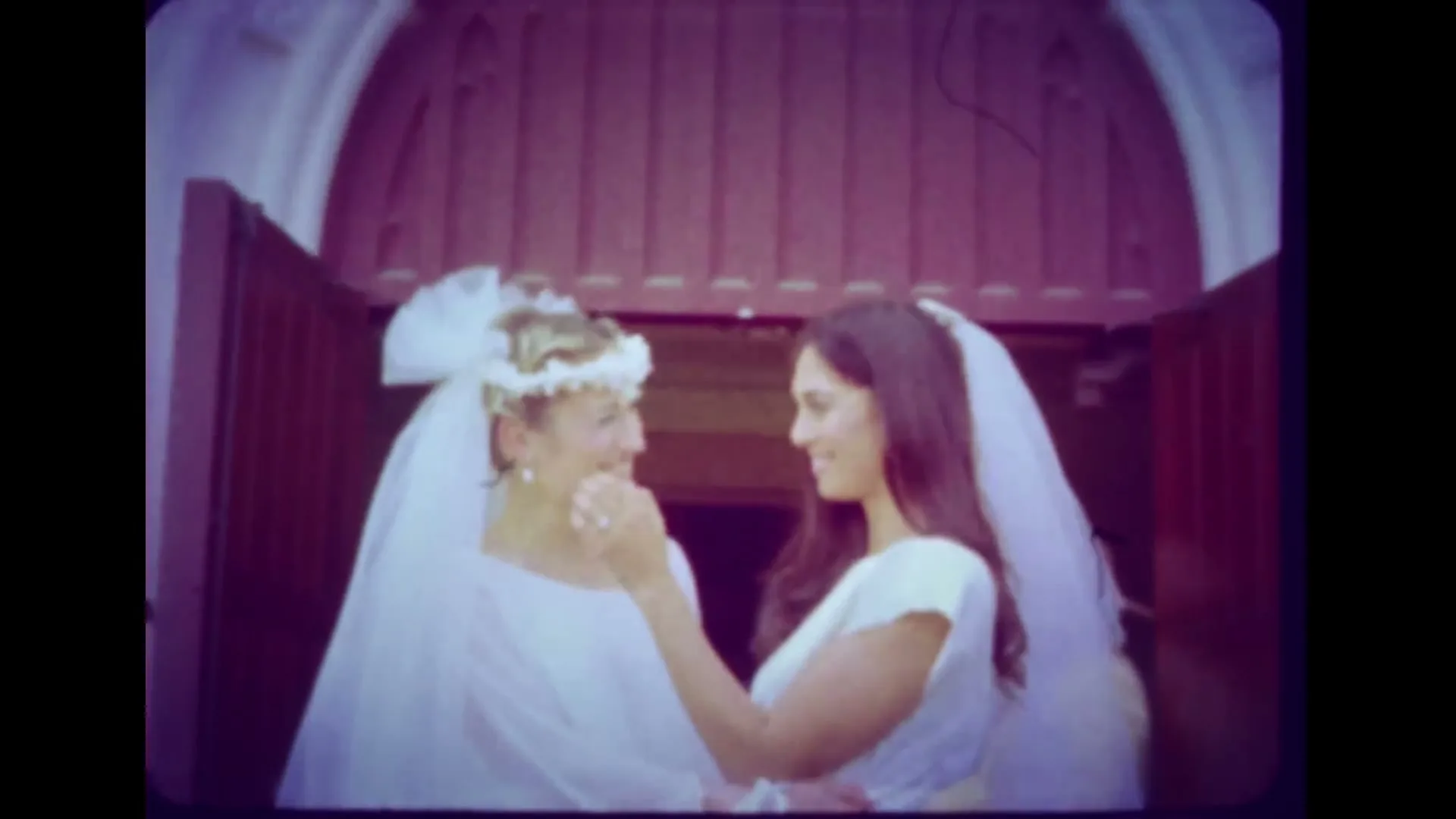 Two women in wedding dresses kiss and embrace in front of a church doorway, captured in a vintage home video style.