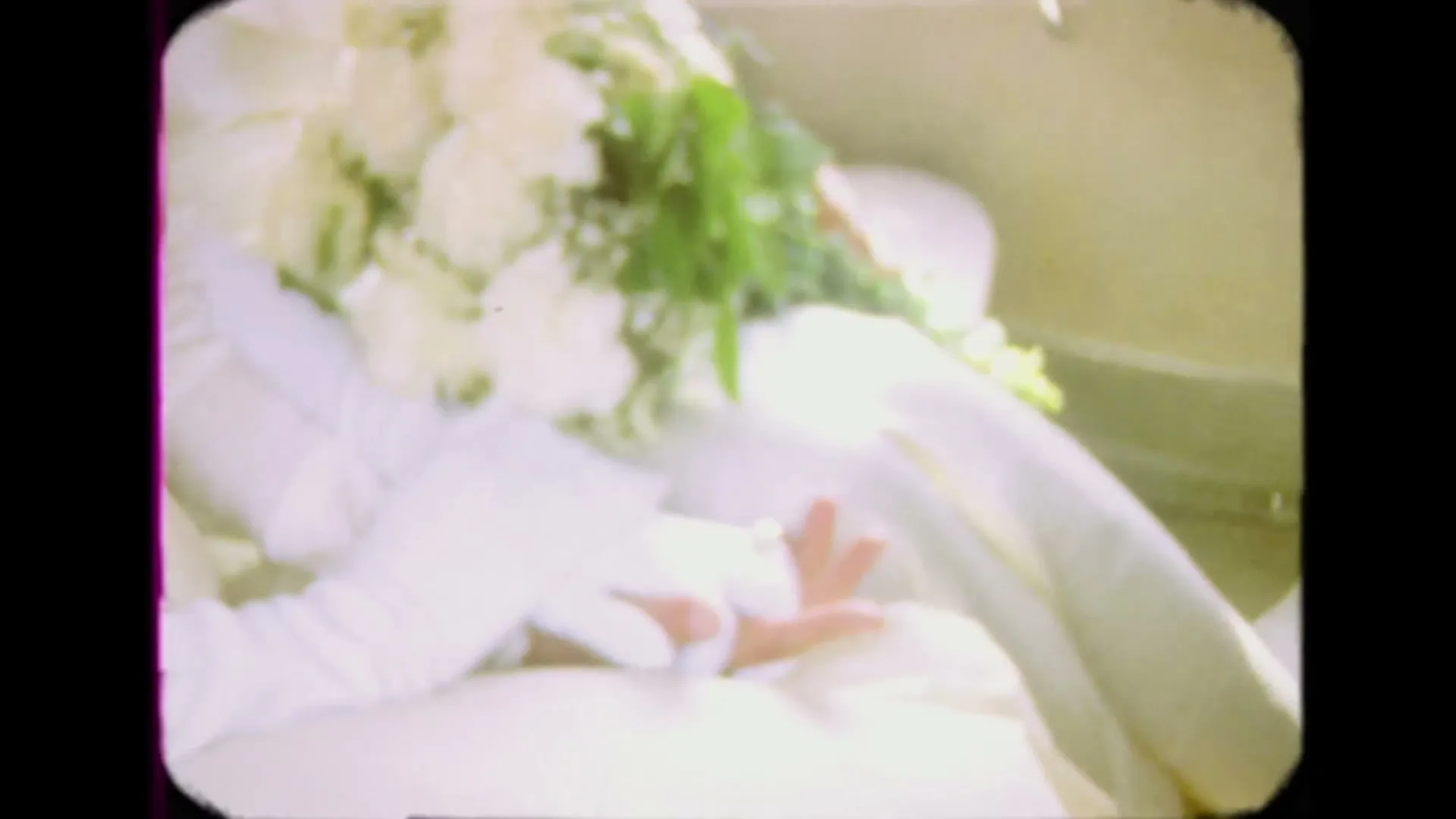 A bride in a white dress and gloves holds a white floral bouquet in a vintage home video shot.