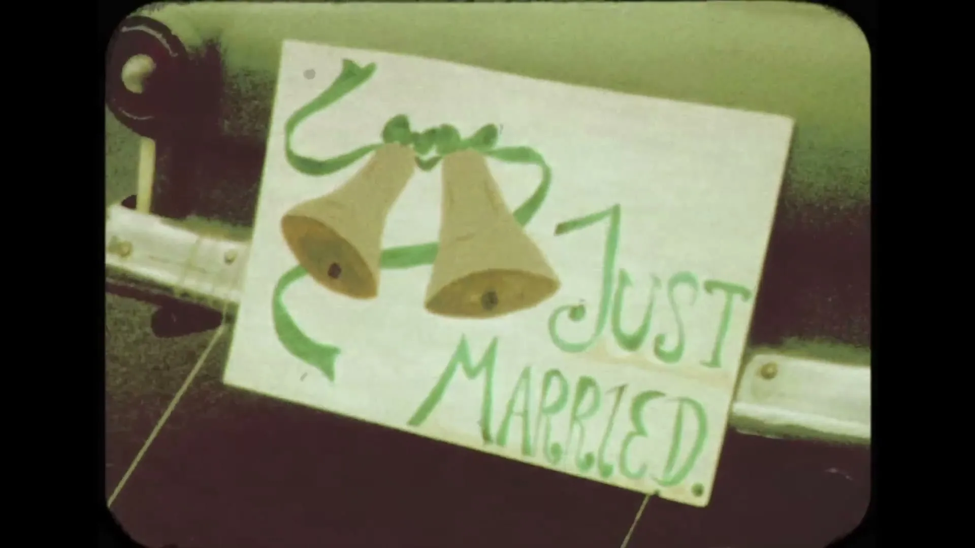 A "Just Married" sign with two bells and green ribbons is tied to the back of a car, filmed with a vintage aesthetic.