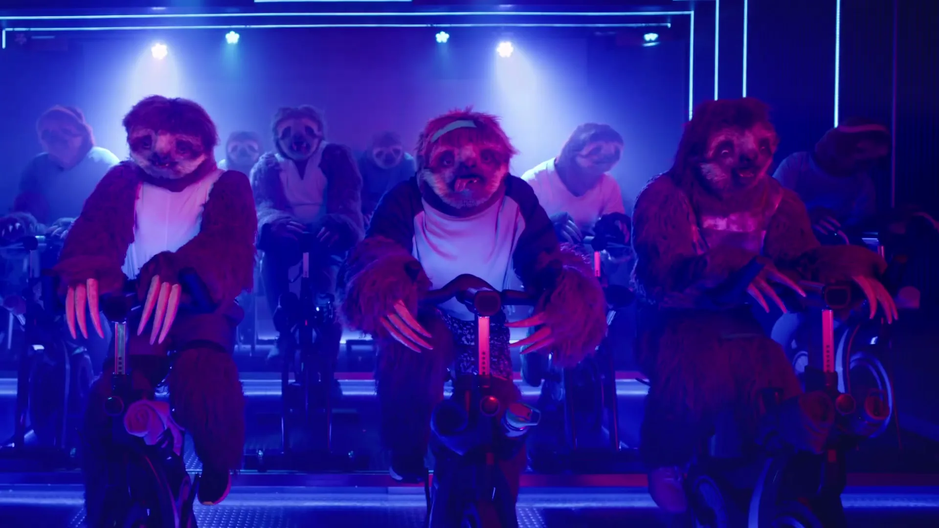 A group of people in sloth costumes are slowly pedaling on stationary bikes in a dark, neon-lit spin class studio.