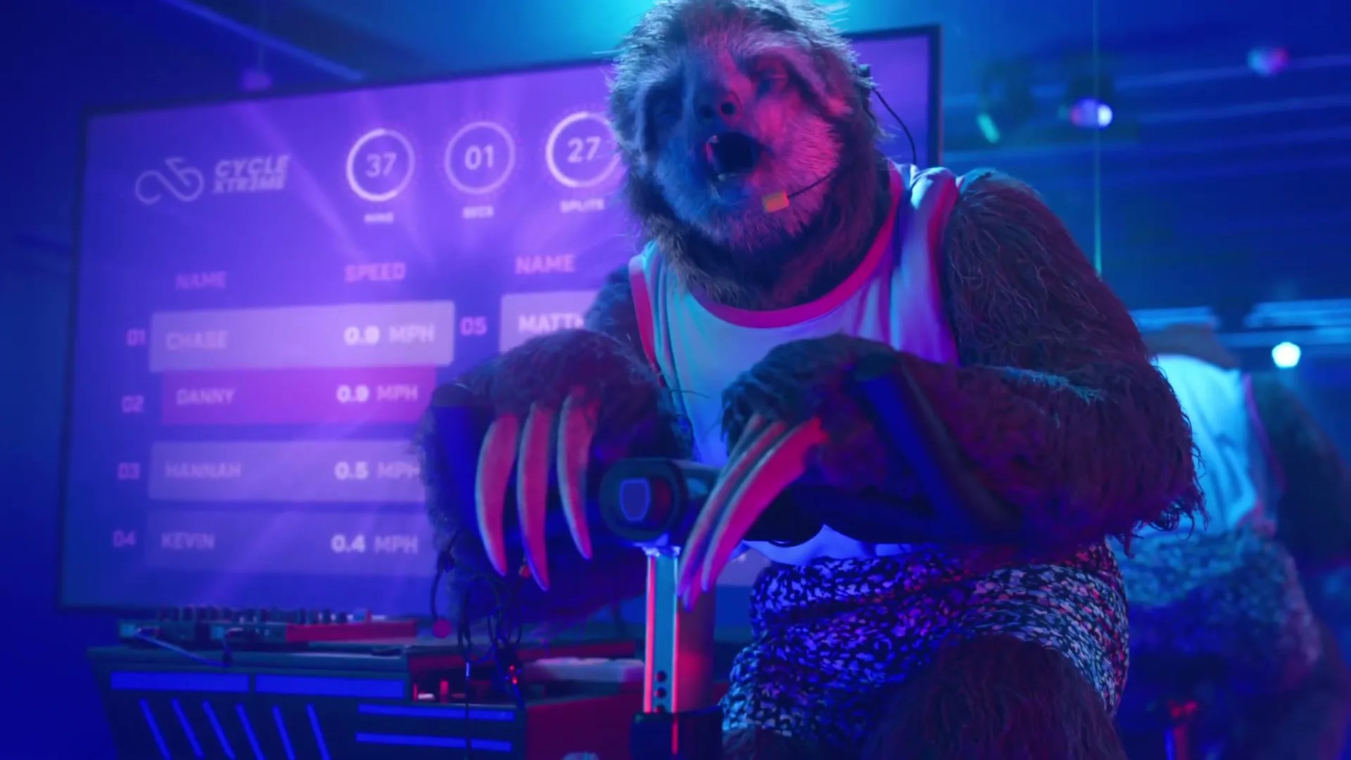A sloth in a tank top and shorts struggles to pedal a stationary bike in a neon-lit spin class, with a large screen displaying low stats behind it.
