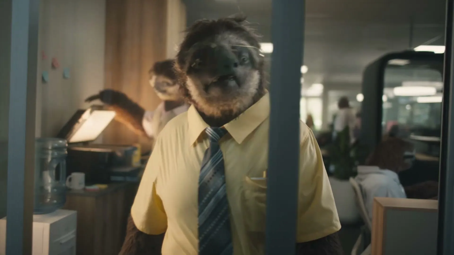 A sloth wearing a yellow shirt and tie with glasses slowly turns its head in an office setting.