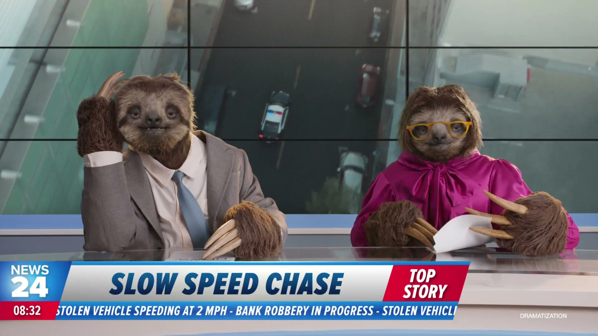 Two sloths dressed as news anchors report on a "slow speed chase" with a police car pursuing a vehicle on a highway shown on a screen behind them.