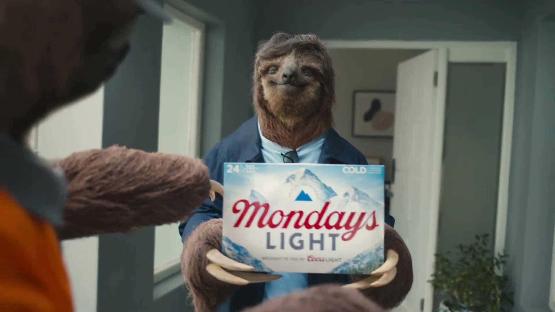 A sloth-like creature in a blue jacket and glasses holds a case of "Mondays Light" beer, smiling at another sloth-like creature in an orange shirt at a doorway.
