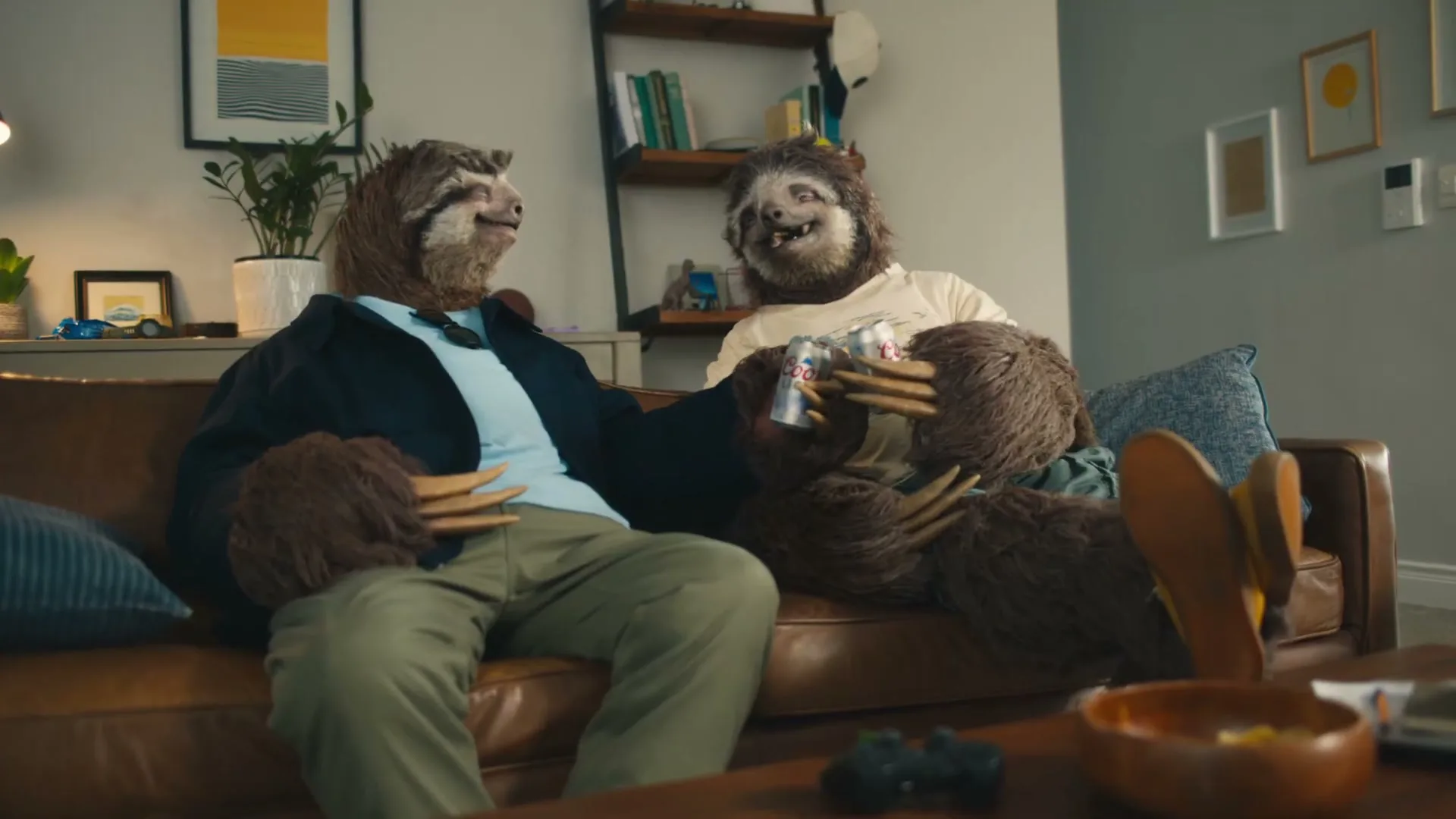 Two sloths in human clothes sit on a brown leather couch, holding Coors Light cans and looking at each other in a living room.