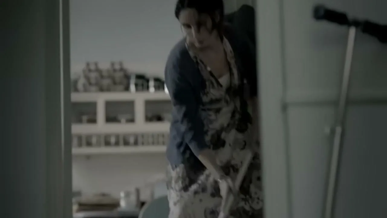 A woman in a dress and cardigan mops the floor in a dimly lit hallway, moving towards a kitchen.