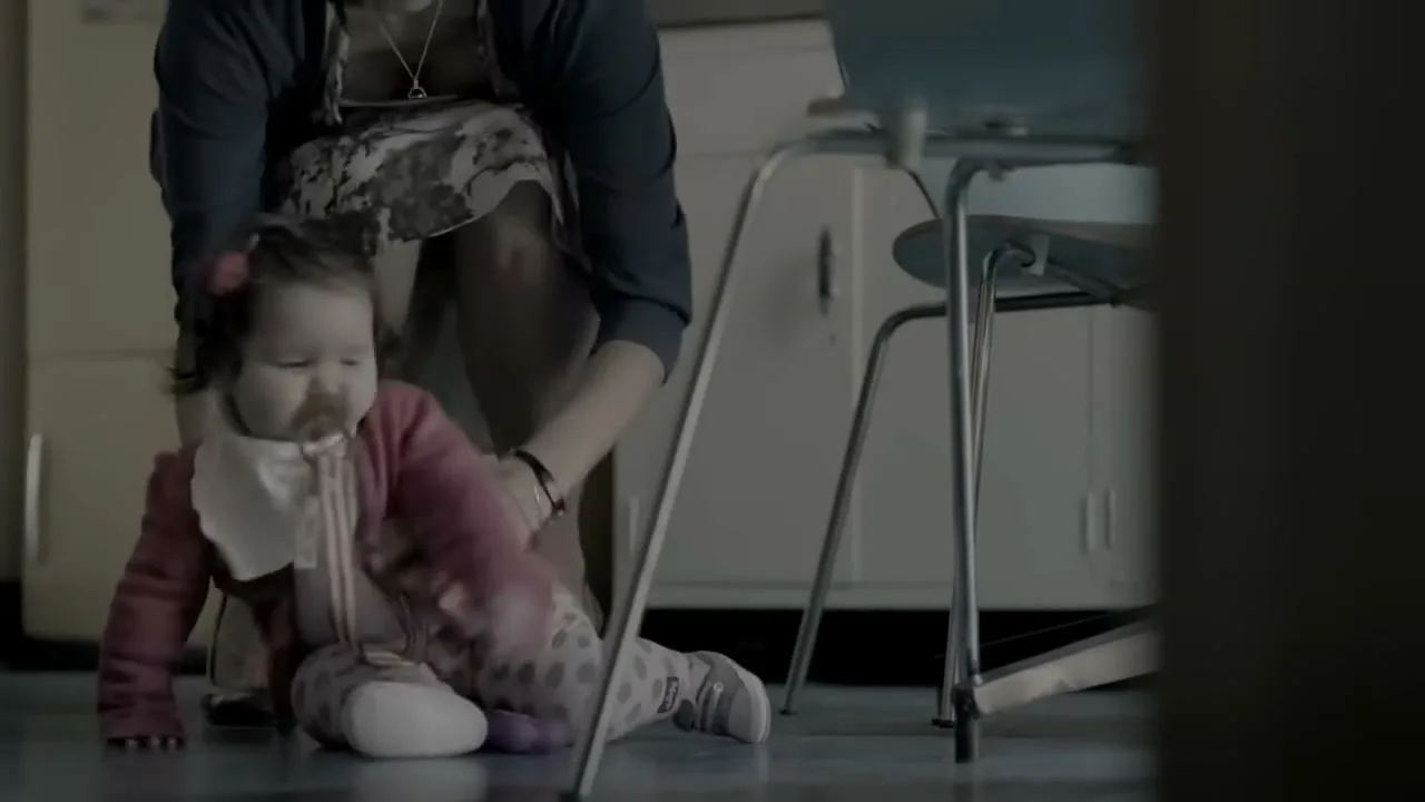 A baby girl with a pacifier sits on the floor while a woman in a dress and heels helps her up in a dimly lit room.