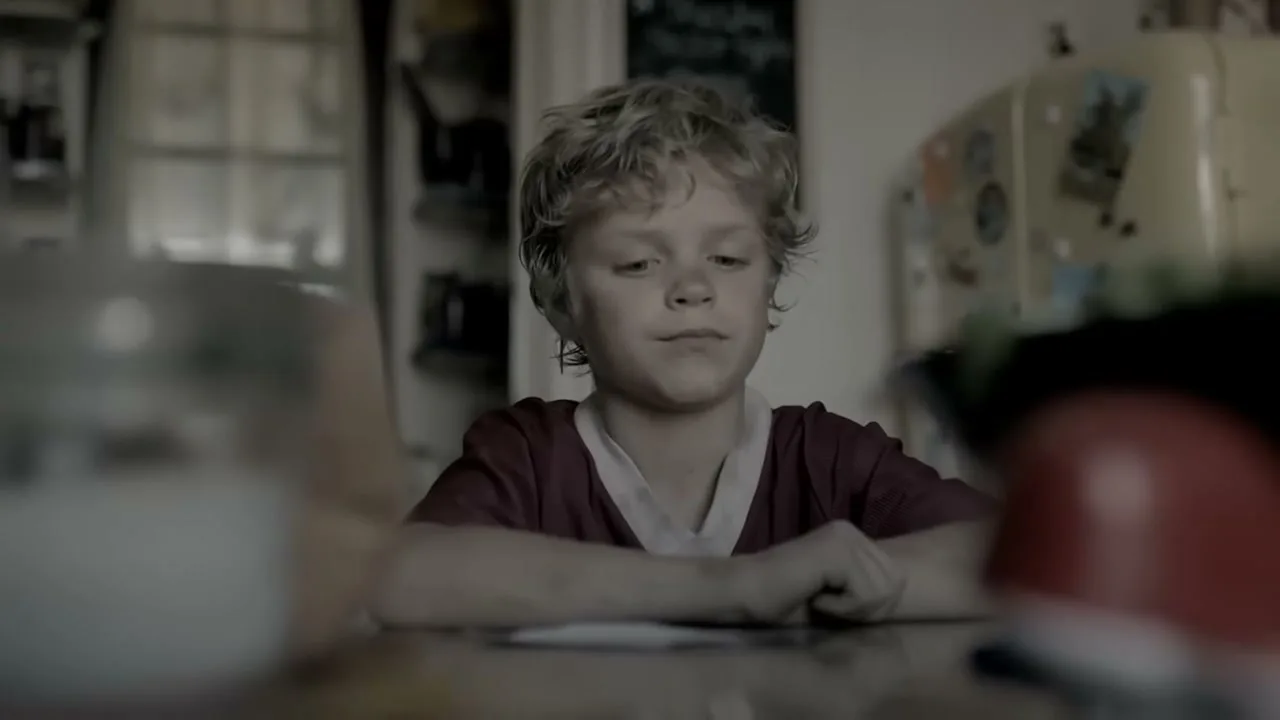 A young boy with blonde hair sits at a table, looking around with a thoughtful expression in a dimly lit room.