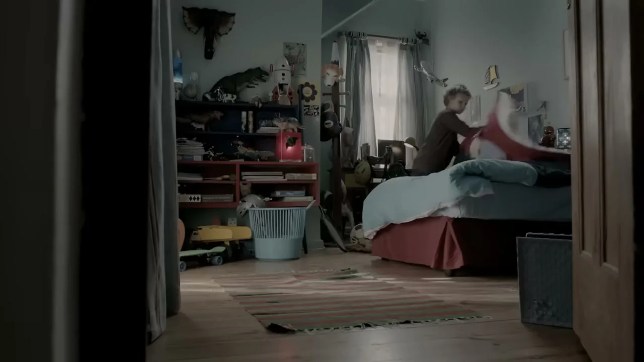 A young boy with curly hair makes his bed in a messy bedroom, seen through a doorway.