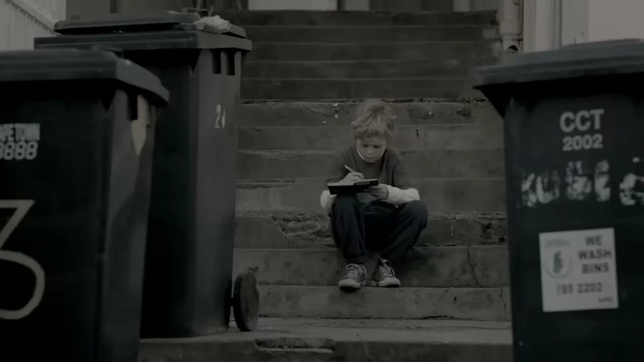 A young boy with blonde hair sits on concrete steps between two trash cans, writing in a small notebook with a pen.