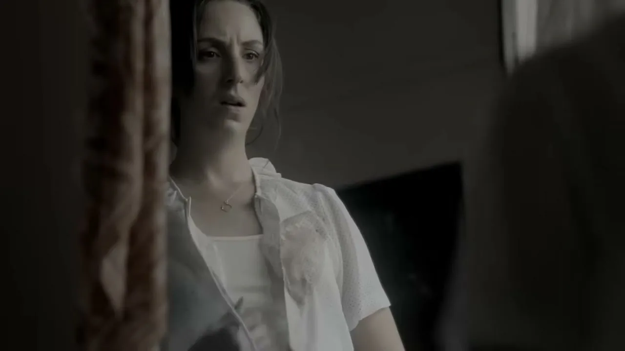 A young woman with dark hair, wearing a white top, looks distressed as she tries to cover a stain on her shirt in an interior setting.